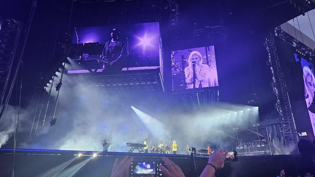 A live concert scene with a band performing on stage, featuring multiple musicians and vocalists. Large screens display close-up images of the performers. The stage is illuminated with lights and surrounded by a misty atmosphere.