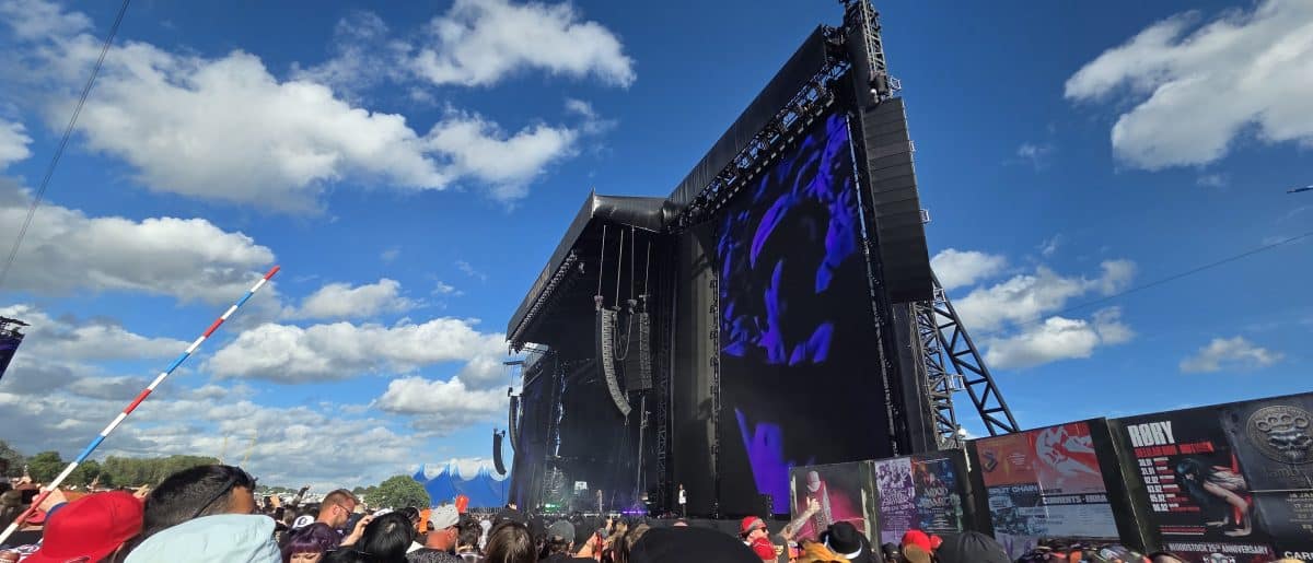 A vibrant outdoor concert scene with a large stage and colorful lighting, surrounded by an audience under a blue sky with clouds.