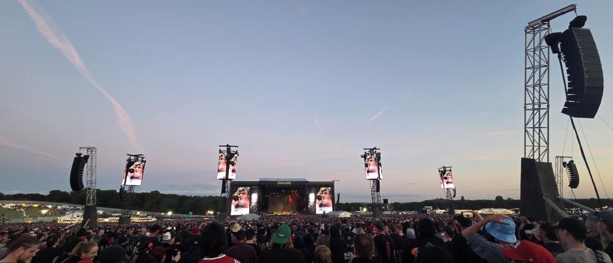 Large crowd at a music festival, with a stage featuring large screens and speakers, under an evening sky.