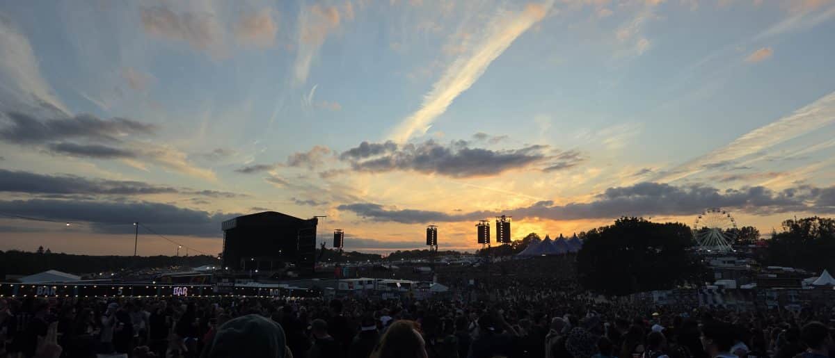A large crowd gathered at a festival during sunset, with a stage and Ferris wheel visible in the background against a colorful sky.
