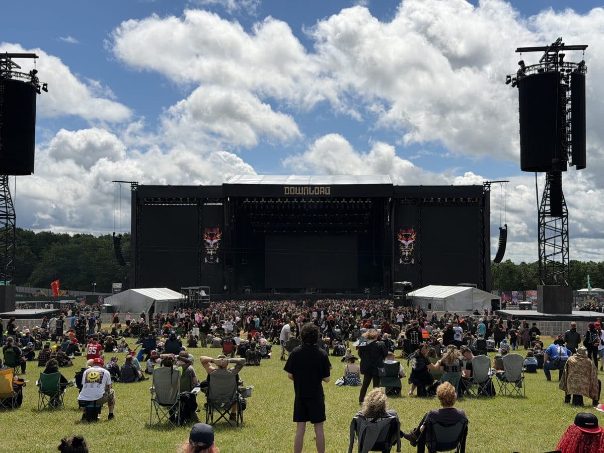 Crowd of concertgoers gathered in front of a large stage during a music festival, with clouds in the sky and a grassy area filled with seated attendees.