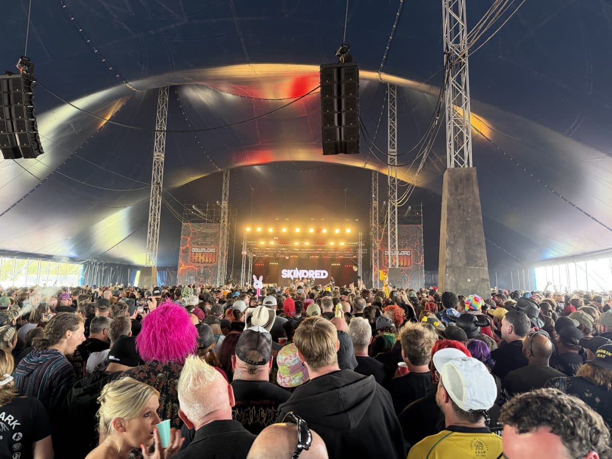 A large crowd of festival-goers gathered under a tent, watching a performance by the band Skindred, with colorful hair and hats visible in the audience.