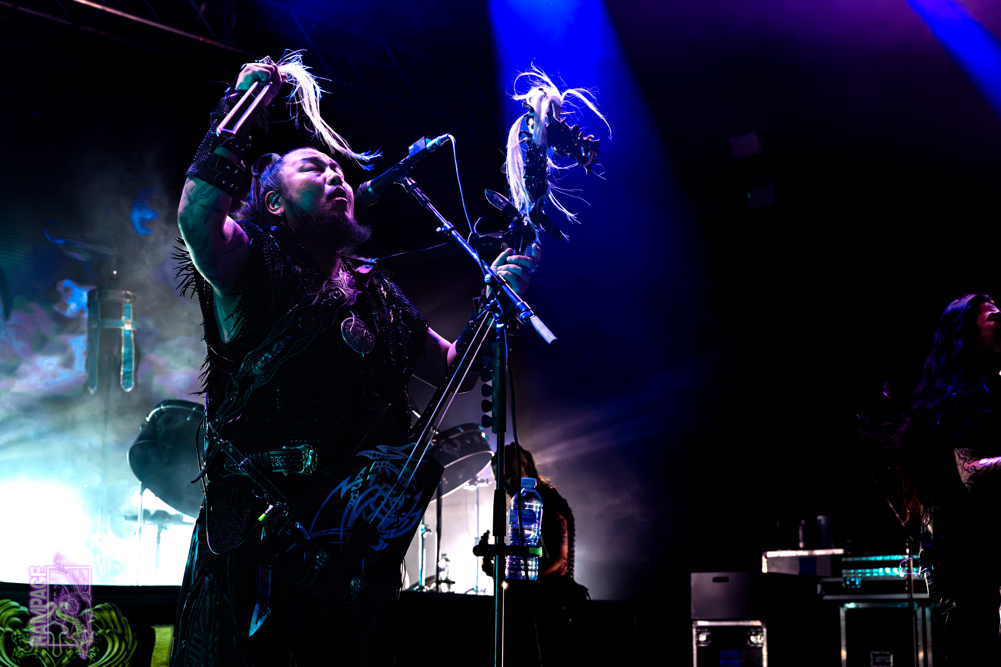A member of The Hu performing on stage, holding the bow of his stringed instrument aloft as he sings into the microphone