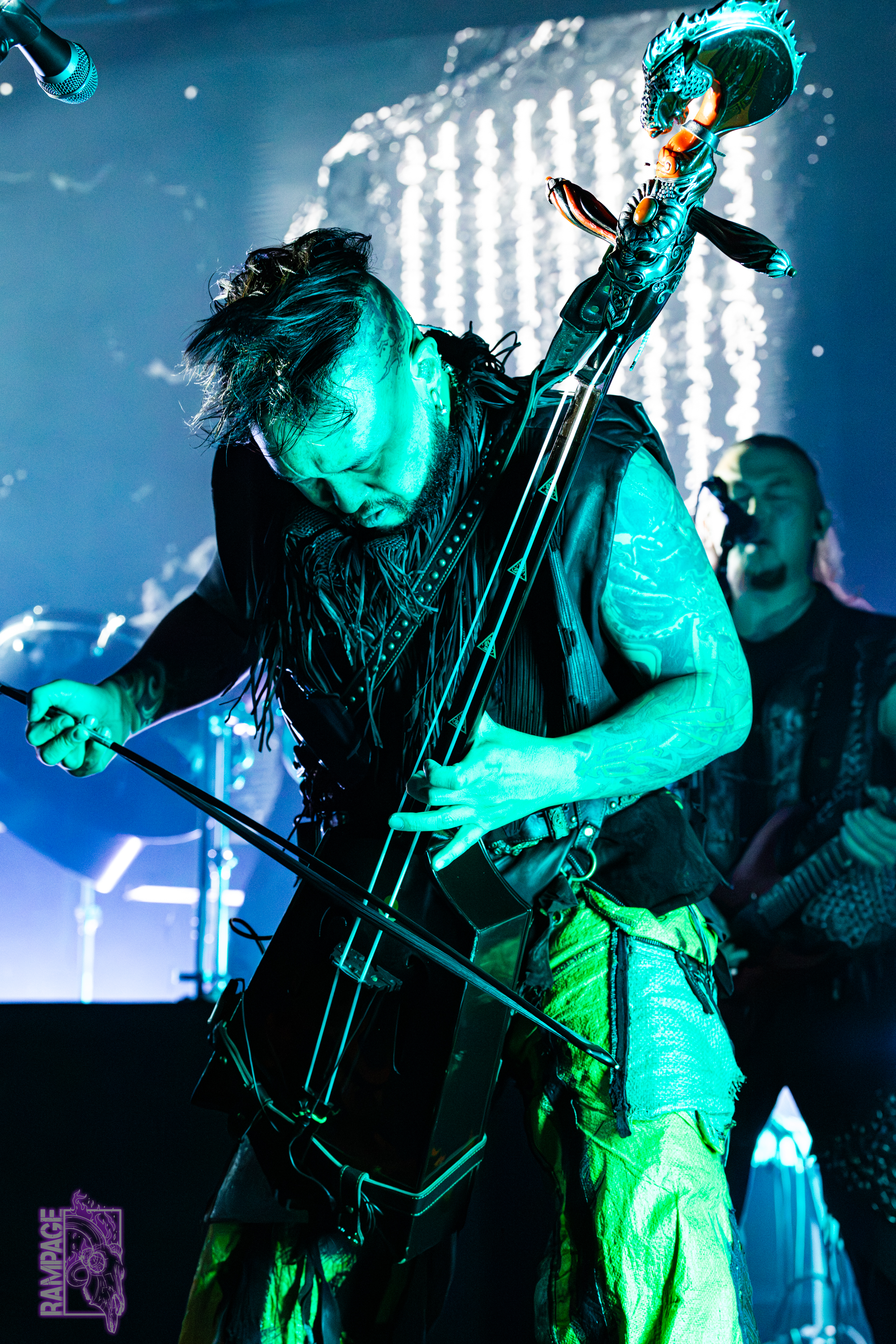 A member of The Hu playing his traditional bowed string instrument, he's looking down at his hands as he plays high up the neck. Behind him, a member of the backing band plays guitar while singing into a microphone